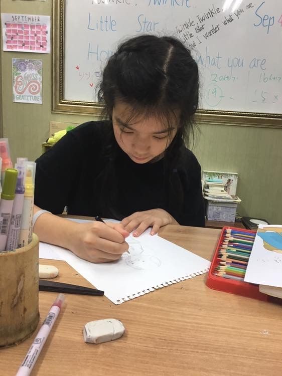 Girl drawing at a desk with colored pencils and stationery in a classroom.