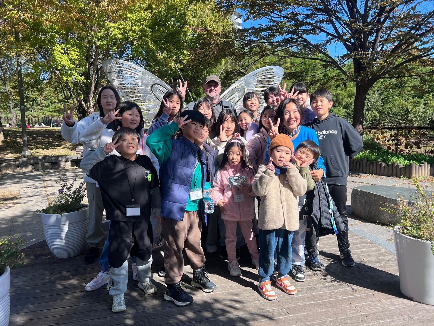 Group of children and adults posing outdoors with trees in the background, making peace signs and smiling.