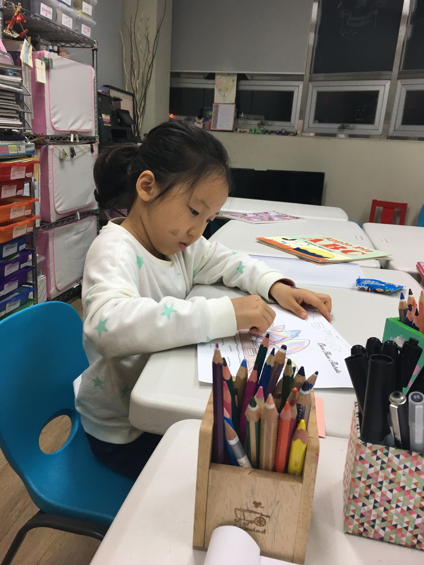 Child sitting at table coloring with colored pencils, surrounded by art supplies in a classroom.