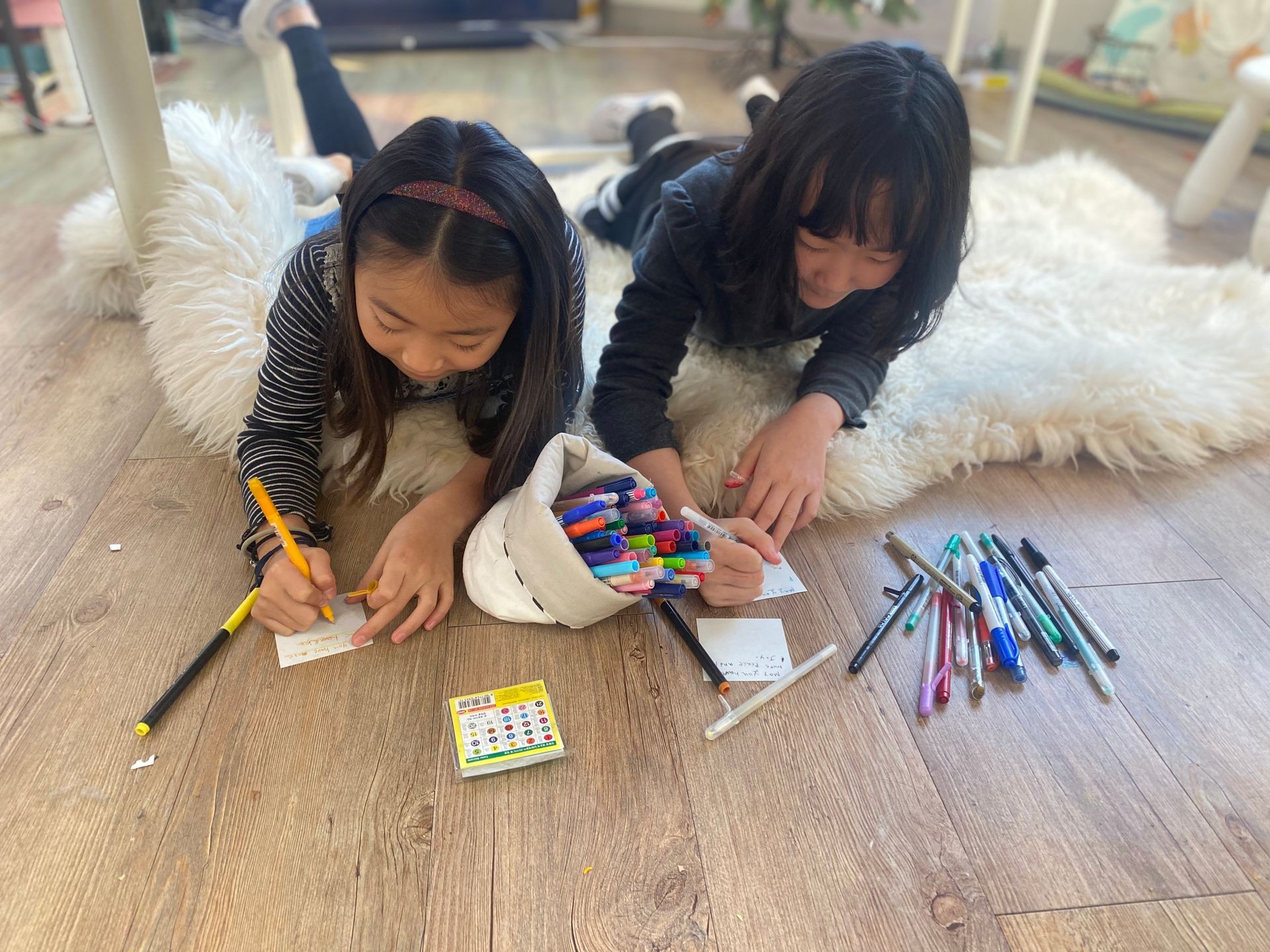 Two children lying on the floor drawing with colored pens on paper cards.