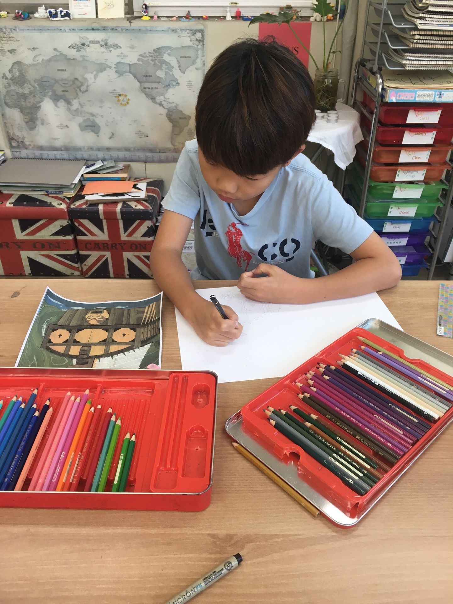 Young boy drawing at a desk with colored pencils and an art book in a classroom.